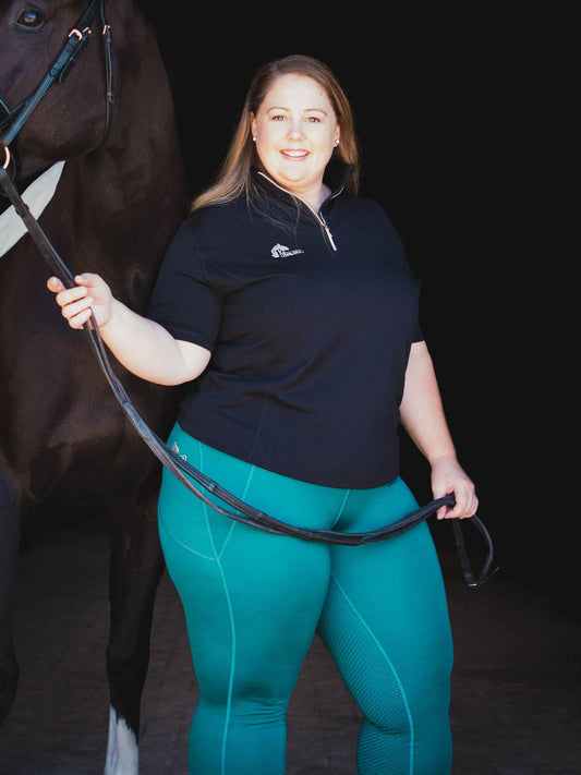 Woman in teal horse riding tights stands smiling next to horse.