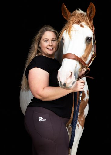 Woman in horse riding tights stands beside a brown and white horse.