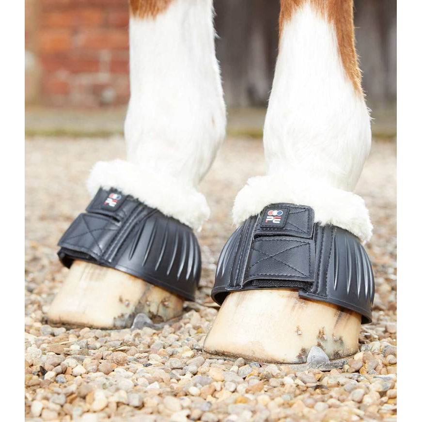 A close-up of a horse's hooves fitted with black protective boots on a gravel surface, with part of its white and brown legs visible.