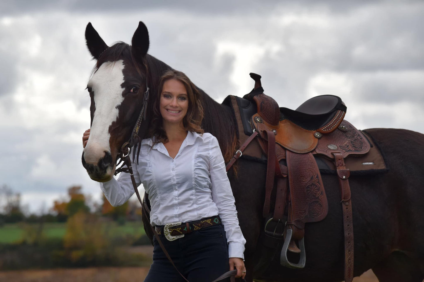 Smiling woman with horse wearing Thinline saddle outdoors, cloudy sky.