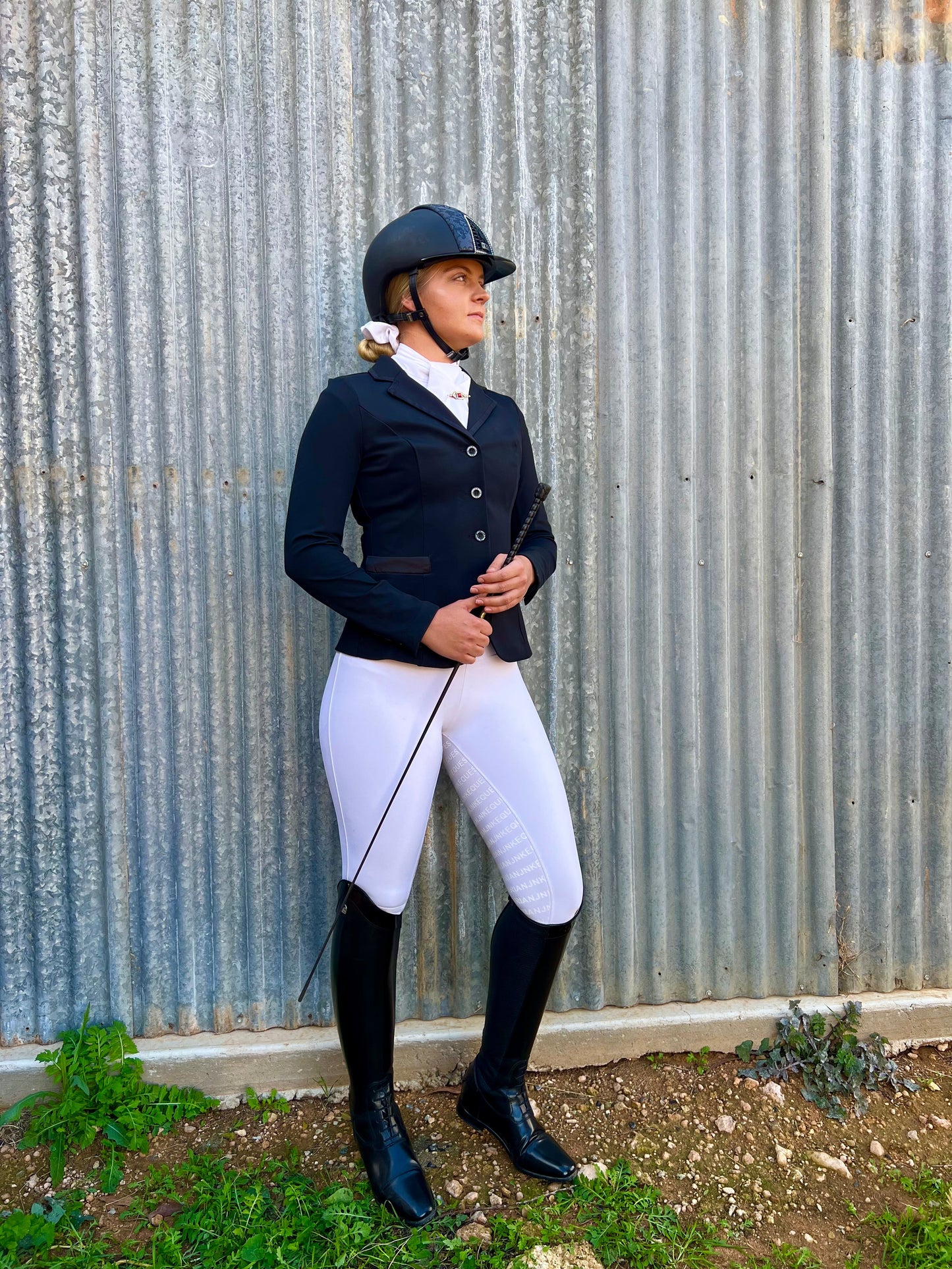 Woman in helmet and horse riding tights stands by corrugated wall.