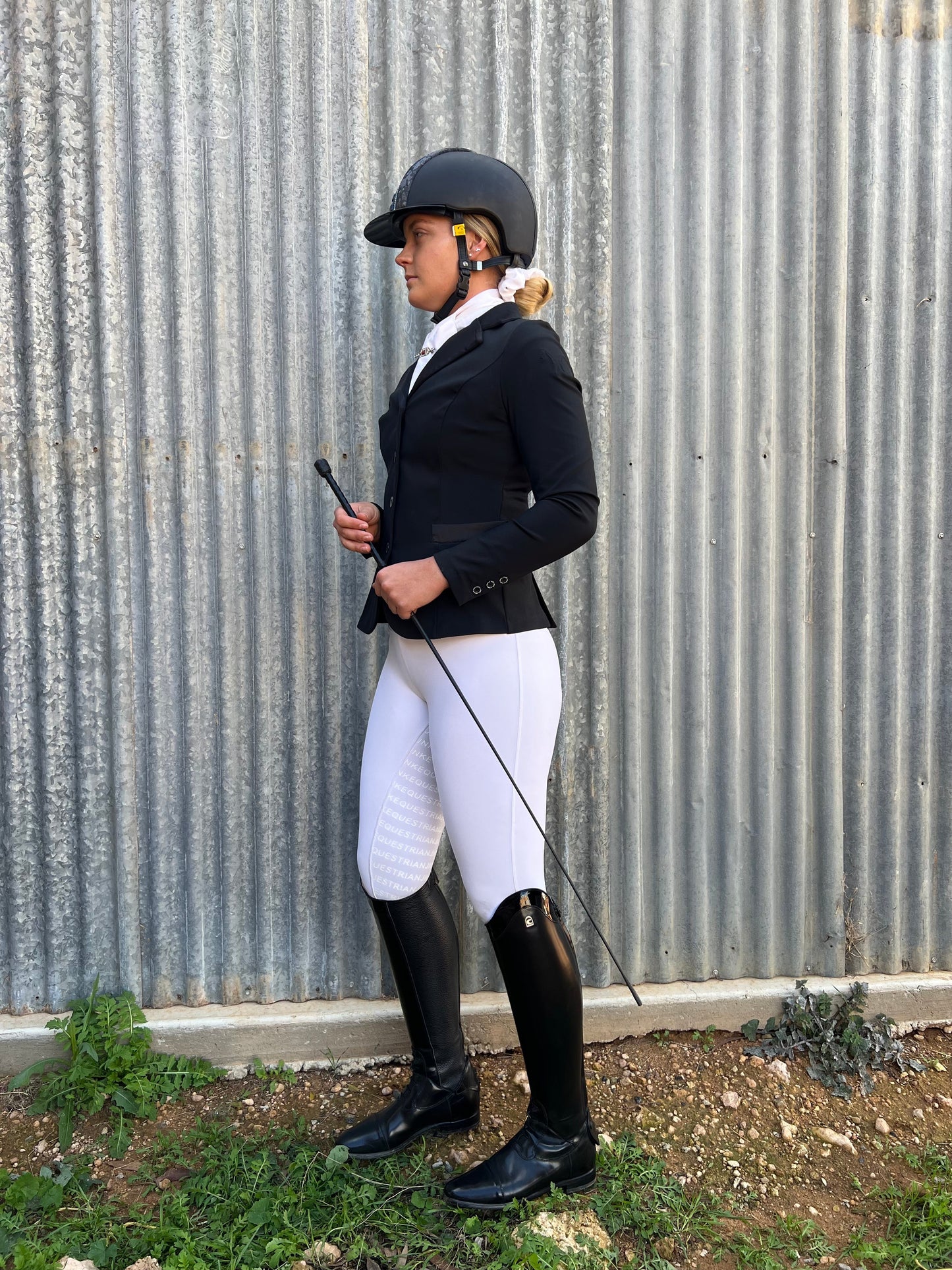 Equestrian in helmet and horse riding tights standing beside metal wall.