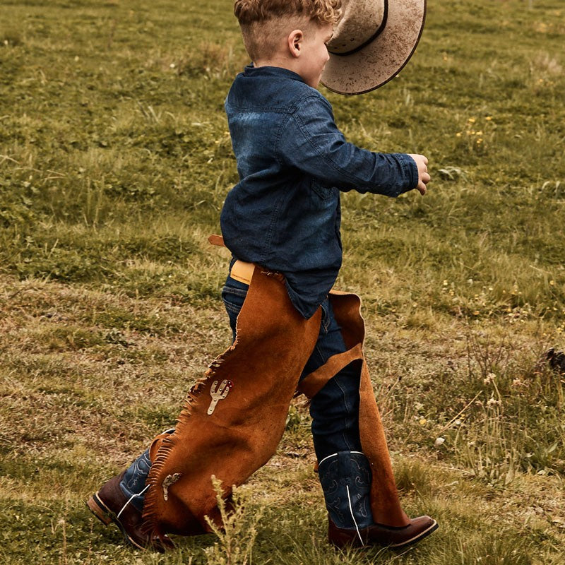 Child in denim wearing Baxter Boots holding hat and chaps outdoors.