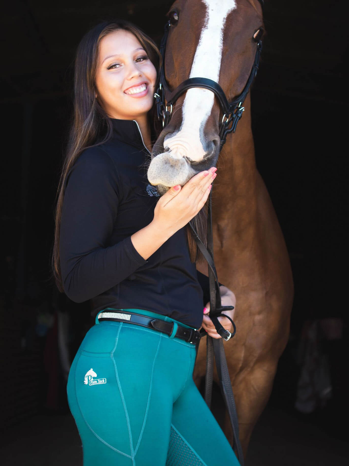 Woman in green horse riding tights smiling with a brown horse.