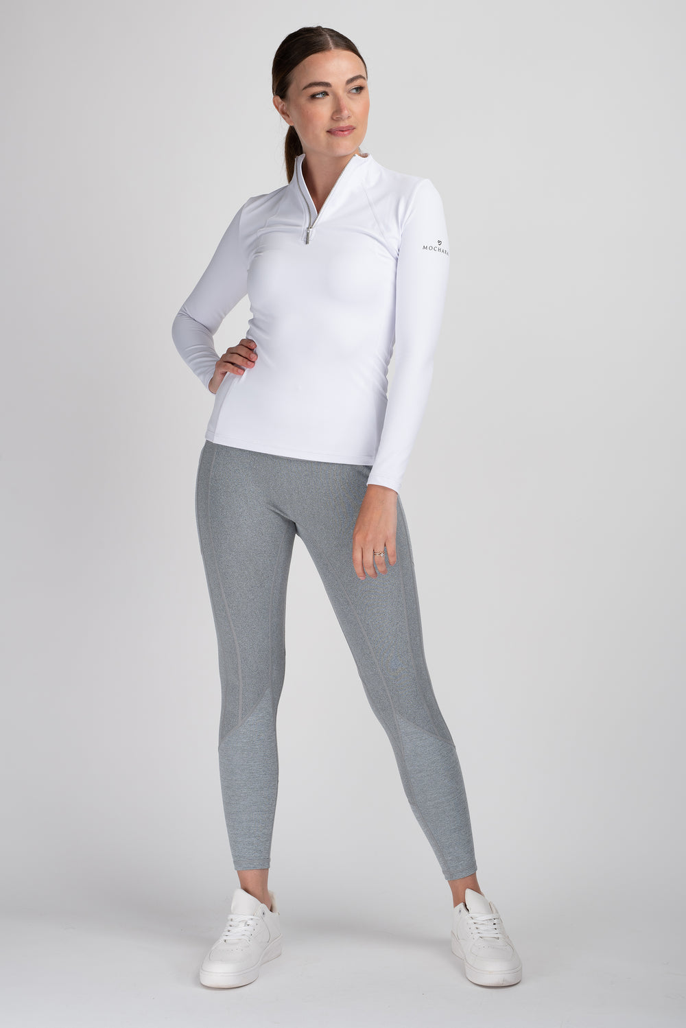 Woman in white top and gray horse riding tights standing confidently.