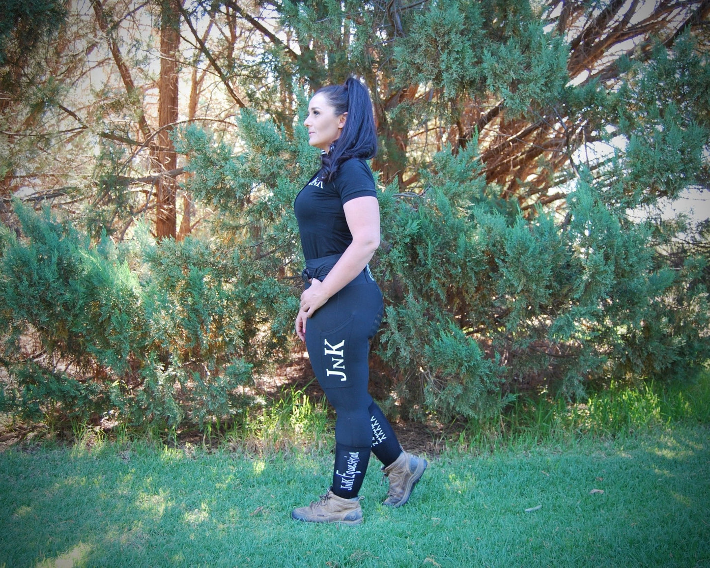 Woman standing in park wearing black horse riding tights and boots.