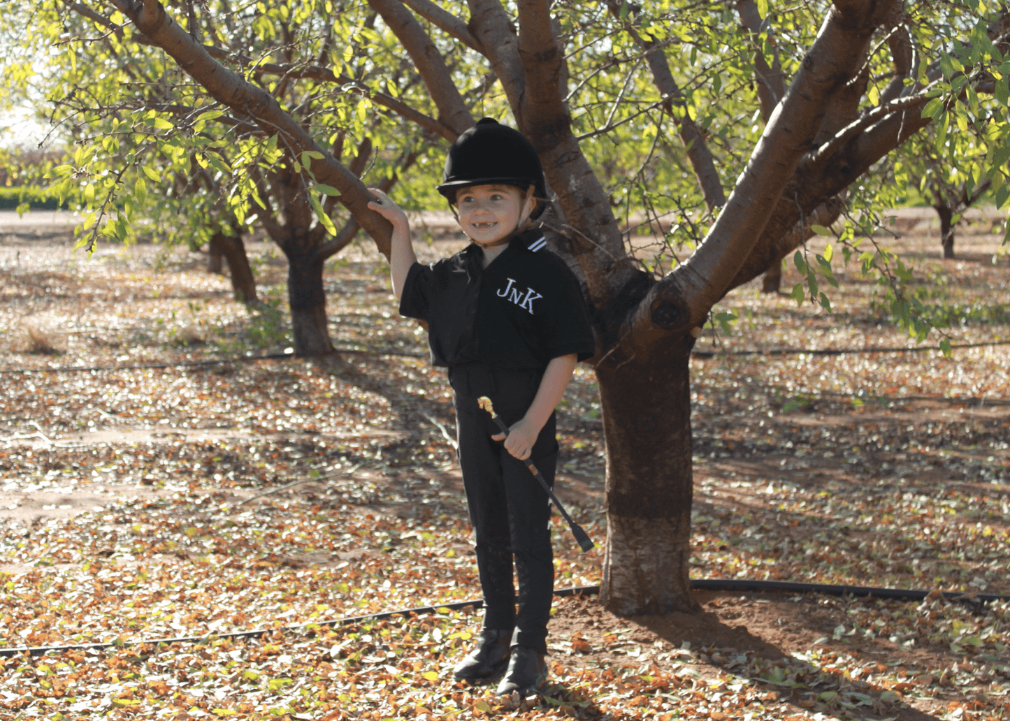 Child in horse riding tights and helmet stands by a tree.