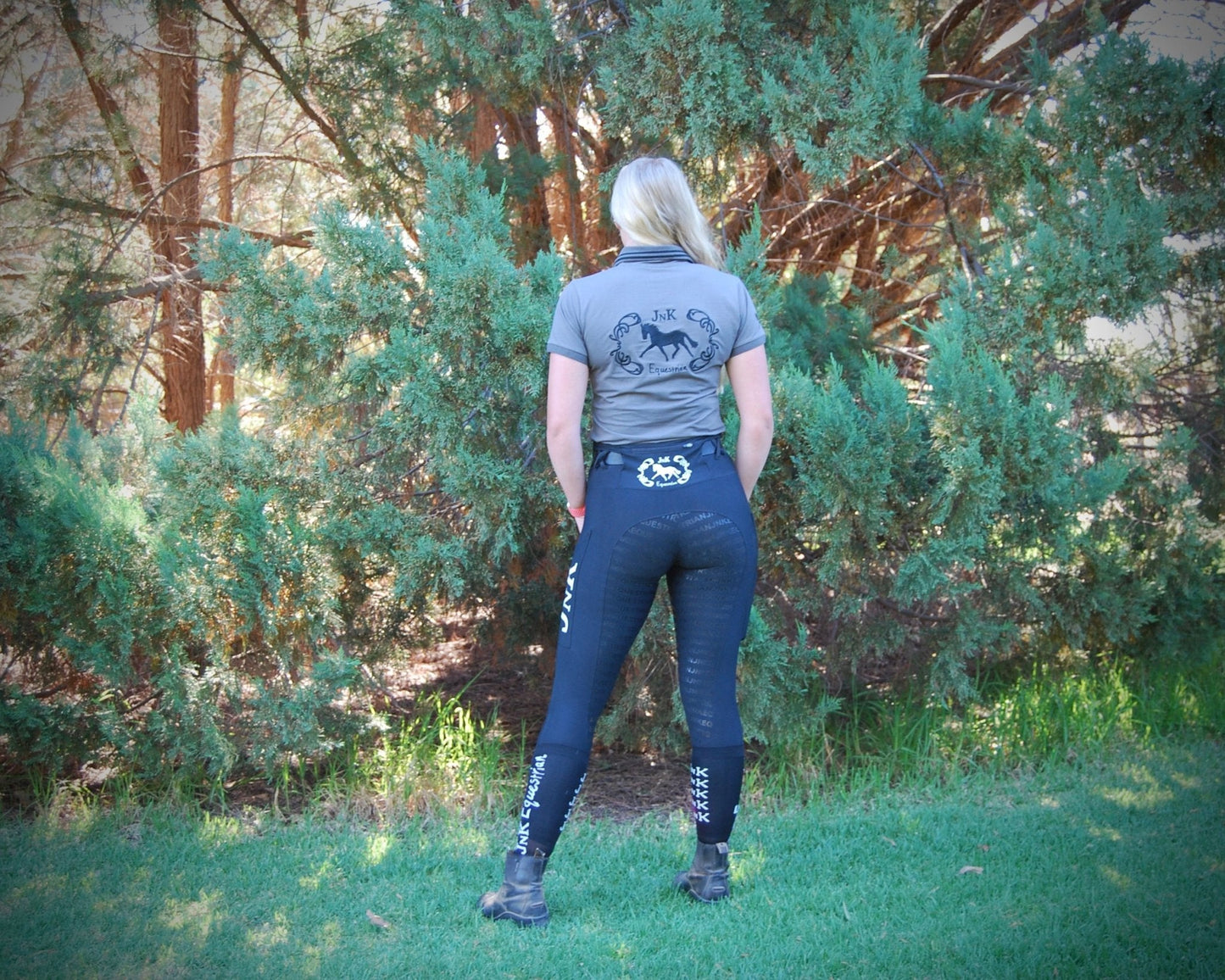Rider in horse riding tights standing outdoors near coniferous trees.