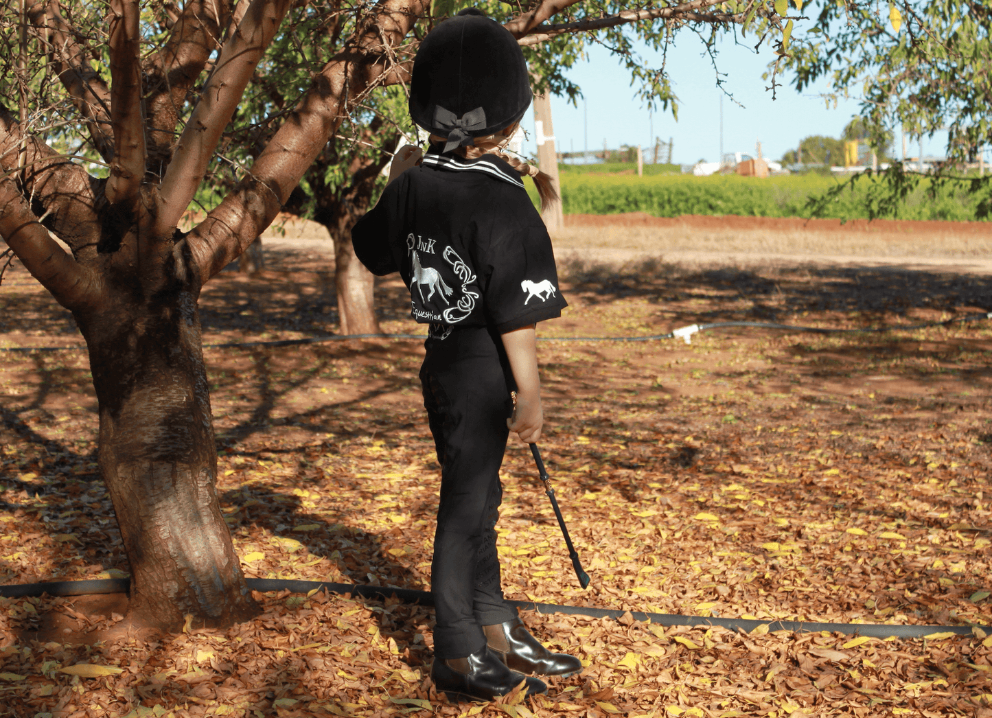 Child in horse riding tights and helmet stands by tree outdoors.