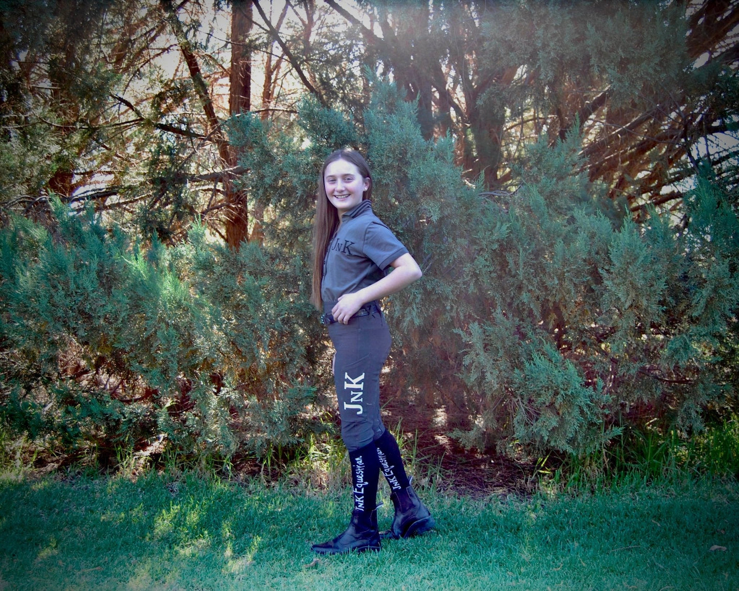 Smiling girl posing in horse riding tights near green bushes.
