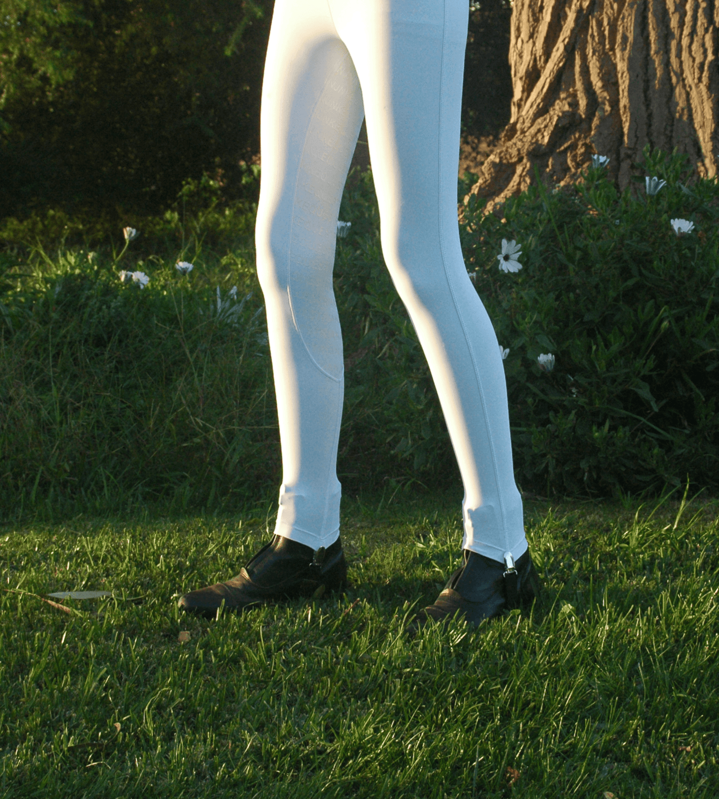 Person in white horse riding tights standing in green grass outdoors.